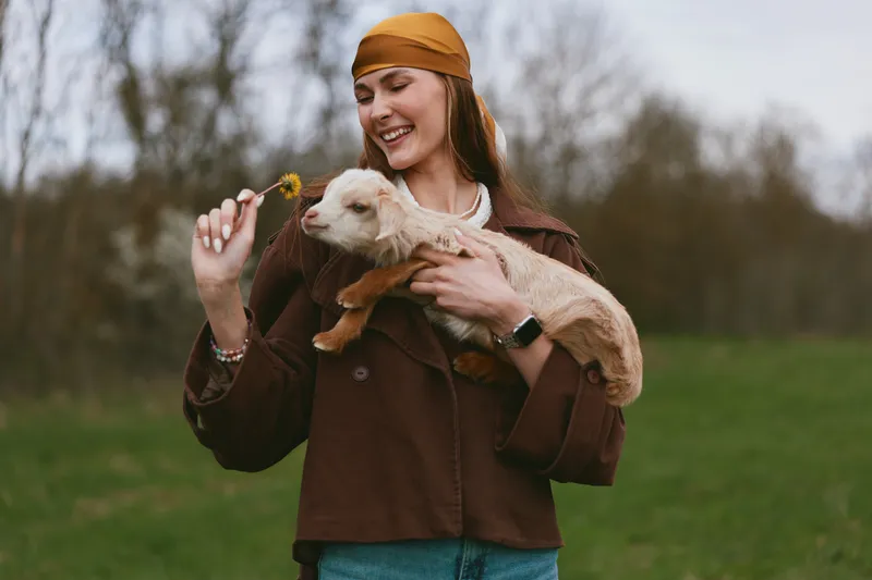 Joyful woman outdoors with a goat kid, smiling as she offers a flower, enjoying a rustic lifestyle moment, natural light, casual clothing, countryside field backdrop.