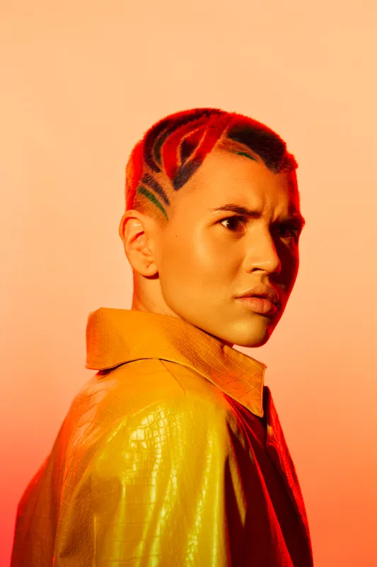 young woman with colorful shaved hair and intense expression wearing yellow jacket against orange background, vibrant style and bold fashion statement