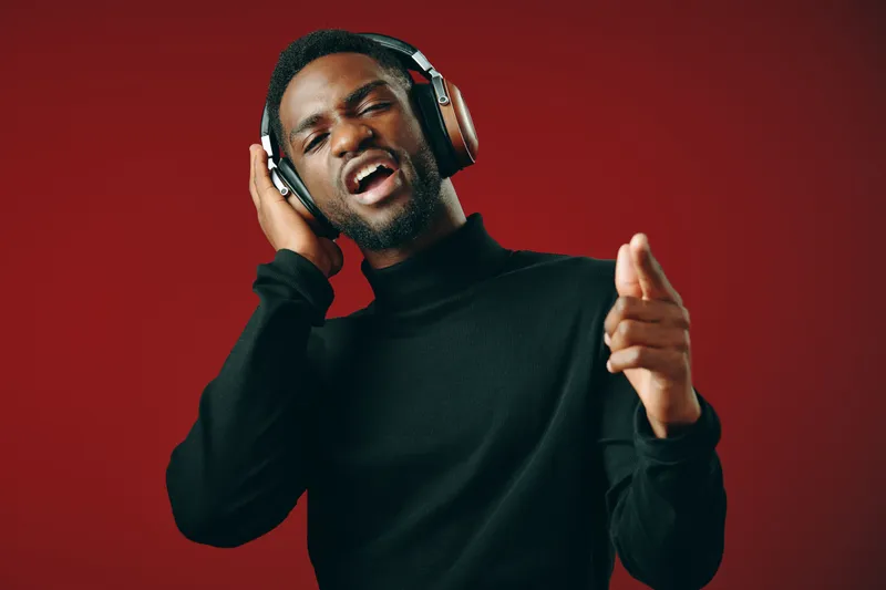 Young Black man singing joyfully with headphones, wearing a black turtleneck, against a vibrant red background, showcasing emotions of happiness and musical enjoyment