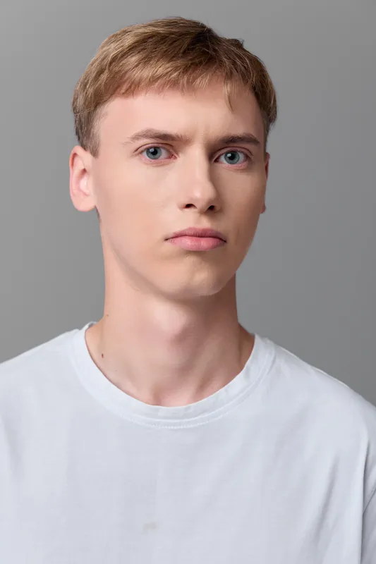 Young man with a neutral expression wearing a white t shirt against a gray studio backdrop, clean lighting and minimalist style suitable for fashion and lifestyle projects