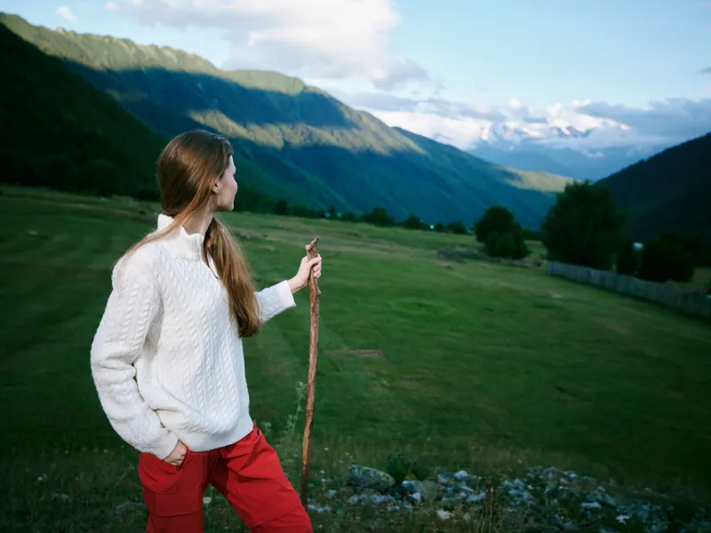 Woman in a white sweater stands with a walking stick in a lush valley, gazing toward distant mountains, an outdoor scene of nature, exploration, and peaceful countryside.