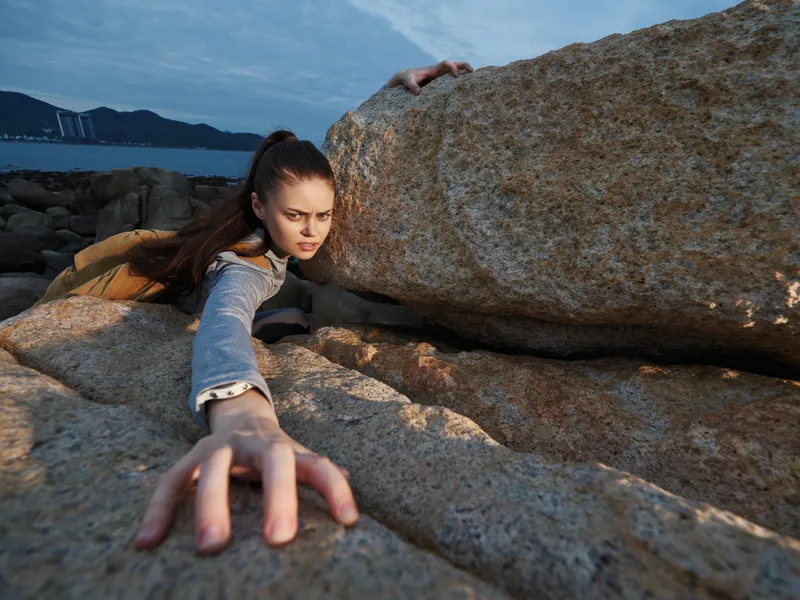 Determined young woman climbing rocks, showcasing strength and resilience in a dramatic outdoor setting with rugged stones and a vibrant sky