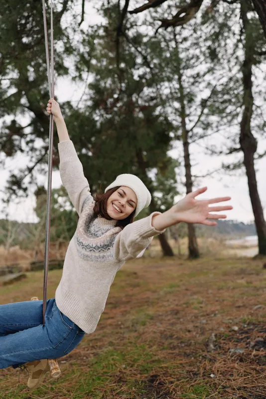 Woman on a swing in a park, smiling and reaching forward, enjoying outdoor leisure. Cozy knit sweater and beanie, trees in the background, autumn light and a carefree moment.