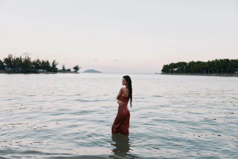 Woman standing waist-deep in calm sea water at sunset, wearing a long red dress, backlit by soft light, long hair flowing, creating a serene coastal portrait with gentle ripples.