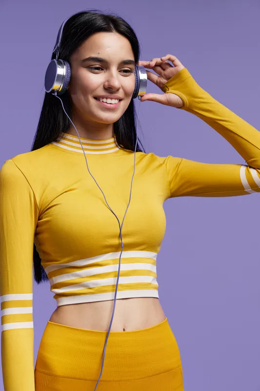 Sport fitness woman wearing a yellow workout outfit and headphones, smiling with long black hair against a purple background in a bright studio setting.