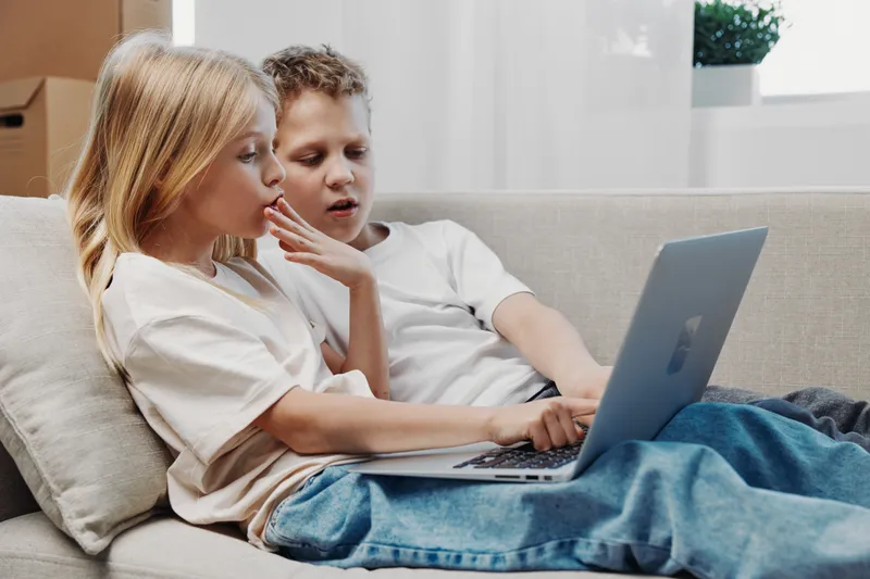 Two children sitting on a sofa using laptop together, focused on screen in a cozy living room setting with natural light and casual clothing.