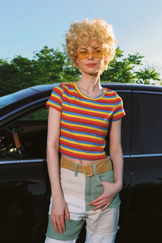 Retro fashion woman lifestyle portrait featuring a vintage colorful striped top, denim shorts, standing by a car under bright sunlight, energetic casual vibe.