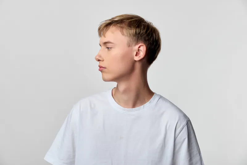 Young man side view wearing a white t shirt in a studio shot, set against a neutral background with a contemplative mood and minimalist styling, head and shoulders framing for a clean, modern look