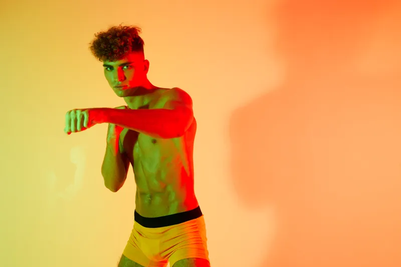 Young man with curly hair posing shirtless in boxing stance against orange background with dramatic red and green lighting and shadow on wall.