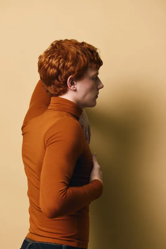 Profile of redhead young man wearing brown turtleneck leaning against beige wall with hand on chest in minimalistic studio setting with soft lighting and calm atmosphere.
