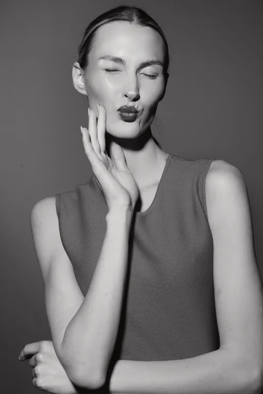 Young slim white woman with pale skin and blonde hair makes a kiss face with closed eyes, posing with hand near cheek in studio. Concept of beauty, expression, and fashion portrait.