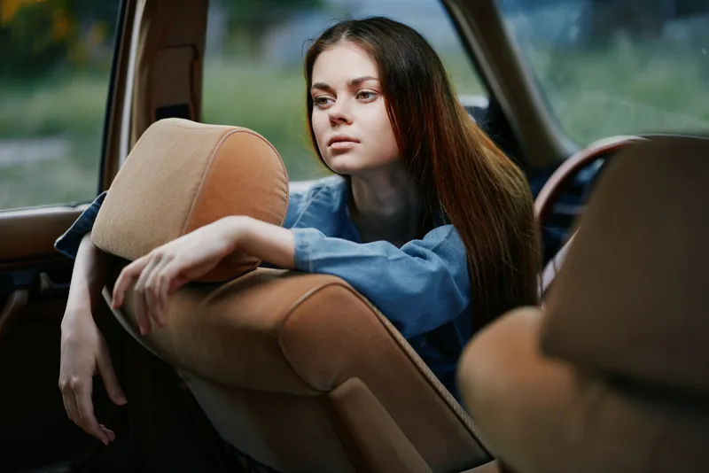 Thoughtful young woman in a vintage car, wearing a relaxed denim outfit, reflecting emotions in a serene natural setting, evoking nostalgia and tranquility.