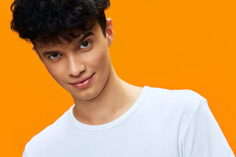 young man in white t-shirt smiling against bright orange background with curly hair and confident facial expression, casual style portrait