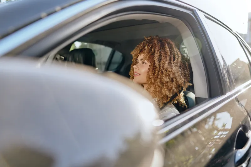 smiling woman with curly hair sitting inside car, looking outside, natural light, casual clothing, urban background, daytime