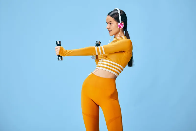 Female fitness enthusiast in vibrant orange activewear lifts small dumbbells against a bright blue studio backdrop, showcasing strength, balance and workout tempo.