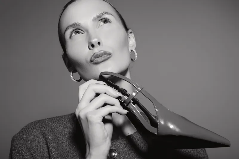 White woman with fair skin holds glossy high heel shoe close to her face, wearing dark textured blazer and hoop earrings. Concept of fashion, elegance, and style in monochrome.