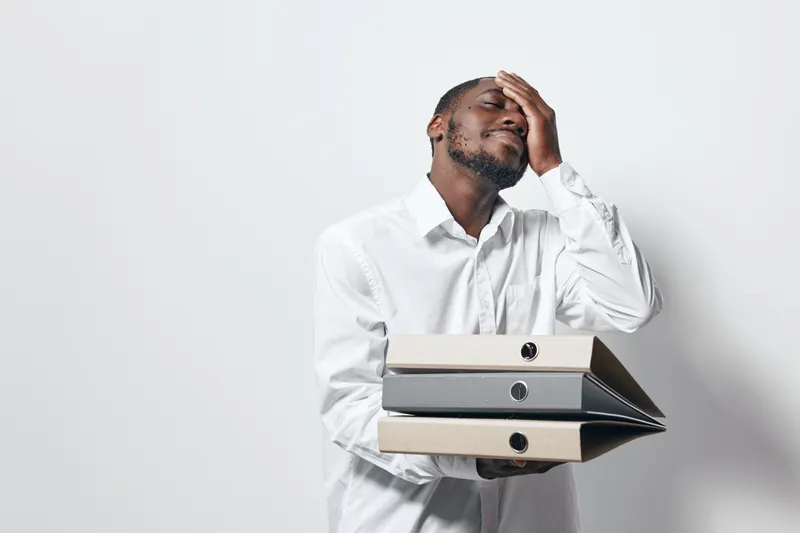 African American man with dark skin holding folders and wearing white shirt, showing stress and fatigue in office environment with plain background.
