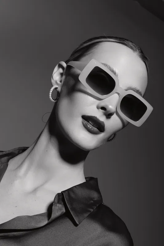 Stylish young white woman with pale skin wearing large sunglasses and hoop earrings poses confidently in studio light. Concept of fashion, accessories, and elegance.