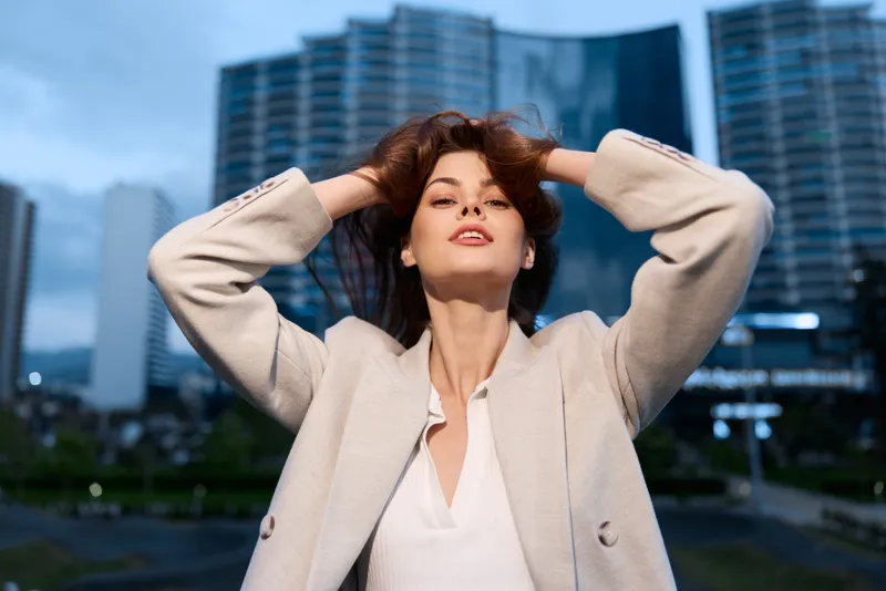 Confident woman in beige blazer posing with hands in hair against modern cityscape at dusk