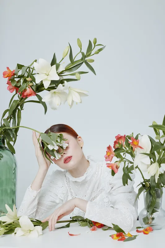 A serene model with auburn hair wearing an elegant white blouse, surrounded by bright flowers in vases, conveying a peaceful vibe in a light colored, minimalist setting