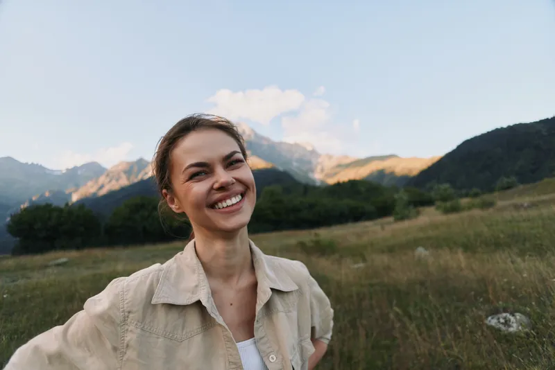 Young woman wearing a beige jacket smiles in a sunlit meadow with mountains in the background, creating a warm, natural lifestyle portrait outdoors, in wide open space
