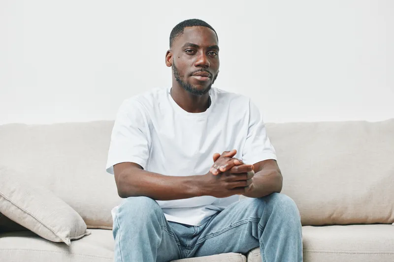 Confident Black man in a white t shirt and jeans sitting on a light gray couch, showcasing a calm demeanor and relaxed atmosphere, ideal for lifestyle content