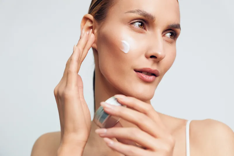 Young woman applying white face cream on cheek with fingers, holding jar in hand. Skincare routine concept with natural makeup and soft studio lighting on white background.