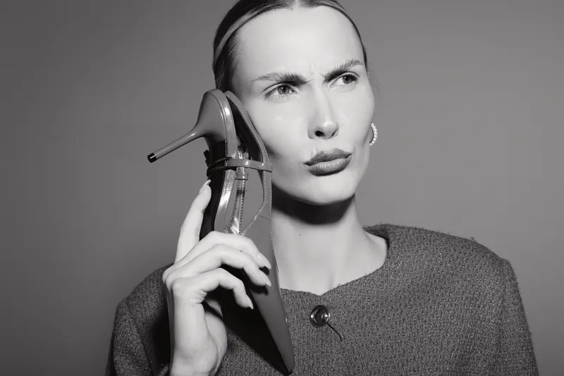 Serious white woman with fair skin holds shoe to ear as if it were phone, isolated on dark background.