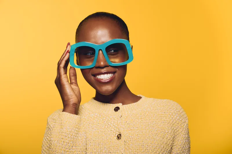 Smiling dark skinned African woman wearing large blue sunglasses and beige sweater poses on yellow background. Concept of fashion, style, confidence, and beauty for black women.