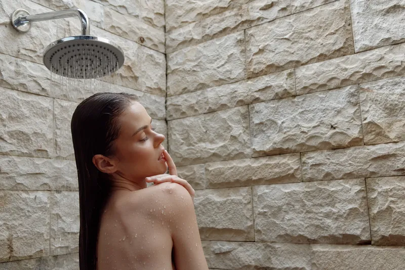 woman showering with wet hair under rainfall showerhead in stone bathroom, natural skin, relaxed mood, wellness and self-care moment