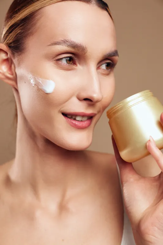 Woman applying skincare cream on face, moisturizer jar in hand, beauty routine closeup, soft spa lighting, fresh clean skin glow, relaxing wellness moment