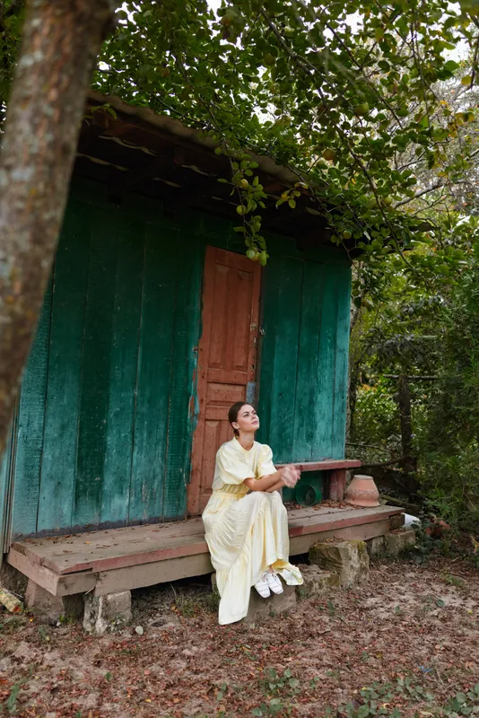 Woman sitting on porch of rustic cottage in rural nature wearing light dress on wooden bench contemplative pose authenticity inclusivity