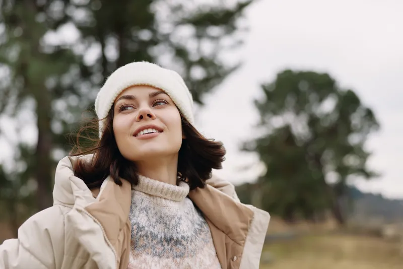 Woman outdoors smiling in a cozy winter jacket and white beanie, standing among trees, soft natural light, relaxed portrait capturing warmth, style and seasonal mood.