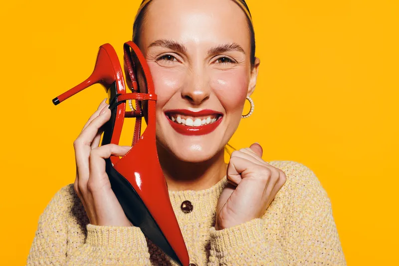 Smiling woman with red lipstick holds red high heel shoes near face on bright yellow background. Concept of fashion, happiness, style, and beauty with vibrant colors and joyful mood.
