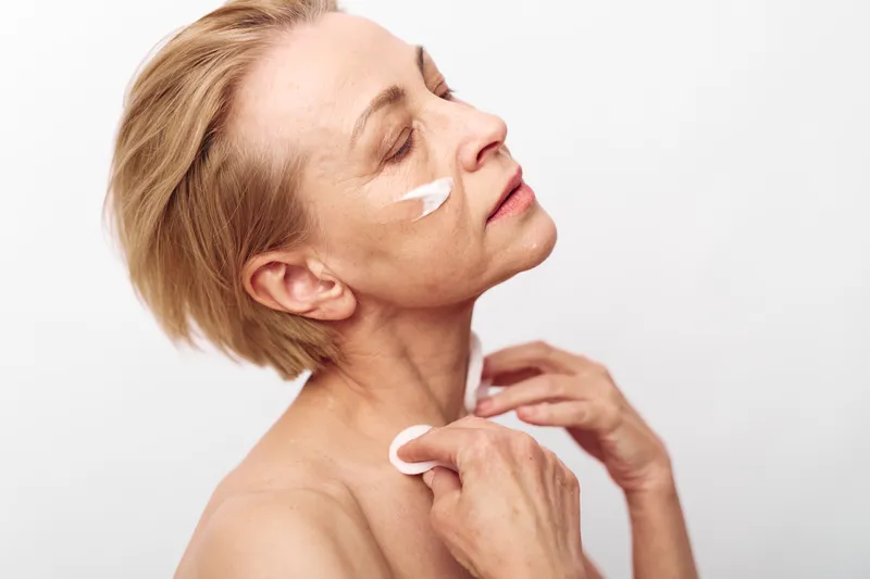 Mature woman applies white body cream to neck and face, showing skincare routine and self-care concept with natural light on plain white background for healthy skin.