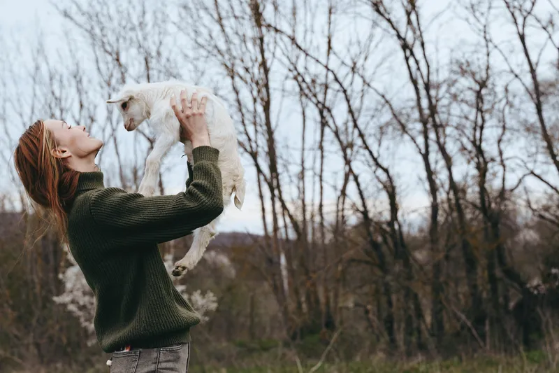 Goat kid is held by a smiling woman outdoors in a rural setting, highlighting lifestyle, affection, and nature. A tender moment, warm light, and joyful bonding with animals create a wholesome vibe.