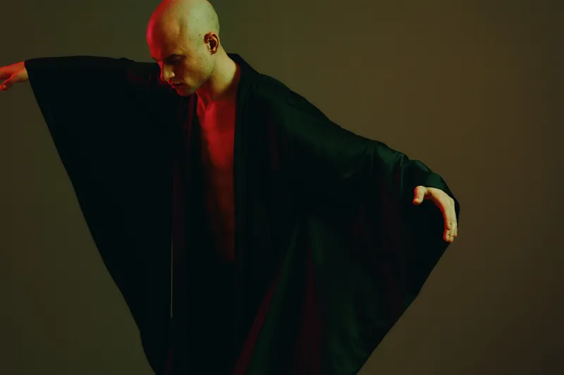 Artistic representation of a man in a flowing black garment, combining elements of movement and grace. The image features bold lighting and minimalistic background, creating depth.