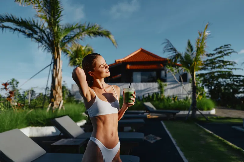 Bikini wearing woman outdoors by the pool enjoys a refreshing drink, radiating vacation relaxation under bright sunshine amid palm trees and a resort atmosphere.