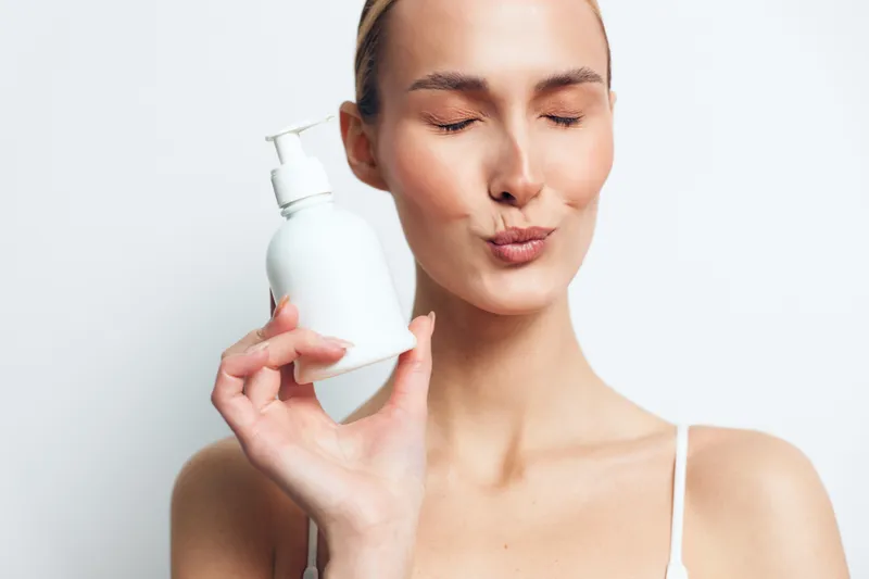 Young woman with clear skin holds a white lotion bottle near her face while making a playful kiss expression against a plain white background.