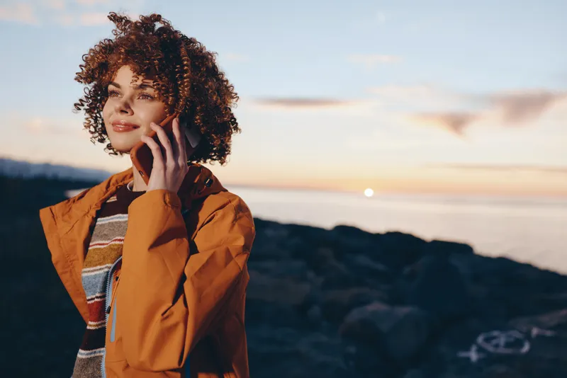 woman lifestyle, sunset coast, curly hair woman holding phone to ear for a call outdoors, near sea and ocean waves, warm golden light, calm relaxed mood