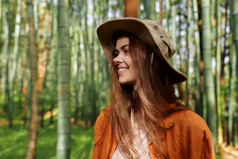 Woman portrait with hat in forest, smile and sunlight on face, nature scene outdoor lifestyle. Brunette model wearing orange coat, candid beauty and peaceful mood in woods.