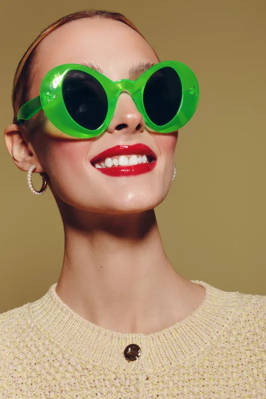 Smiling woman with bright red lipstick wears large green sunglasses and beige sweater. Studio portrait with warm lighting and smooth skin texture. Fashion and beauty concept.