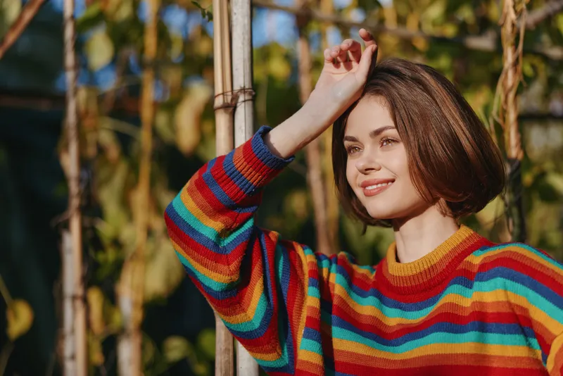 Woman smiling in a colorful striped sweater outdoors, young adult with short brown hair leaning on a bamboo pole in a tropical garden, relaxed lifestyle portrait.
