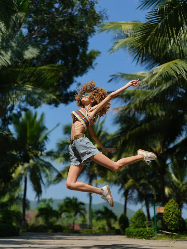 energetic young woman jumping high outdoors, wearing casual summer clothes and sunglasses, surrounded by tall palm trees under clear blue sky