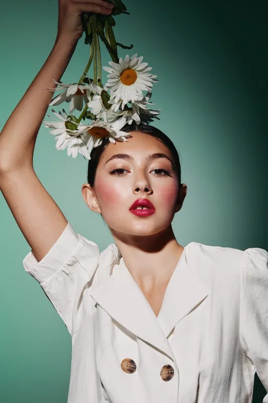 Fashionable woman in white jacket holds daisies above head, showcasing creative beauty makeup with glowing skin, red lips, and pink blush. Studio shot with soft light on green background.