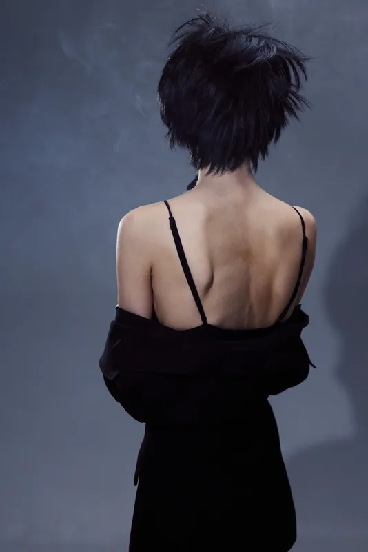 Back view of a young woman with short black hair wearing a black off-shoulder outfit against dark background.