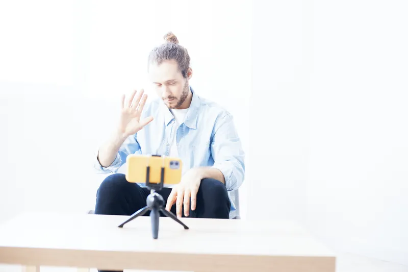 Young man in casual clothing, waving at smartphone camera, captured in a bright, minimalistic interior with soft colors, representing online communication and connection