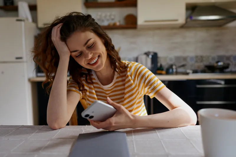Bright kitchen scene with a smiling woman at home, wearing a striped tee, using a smartphone, showcasing a relaxed casual lifestyle and a cozy domestic moment in natural light