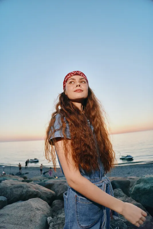 young woman with long curly hair wearing a red bandana and denim outfit enjoying sunset at rocky beach with calm sea and clear sky in warm evening light