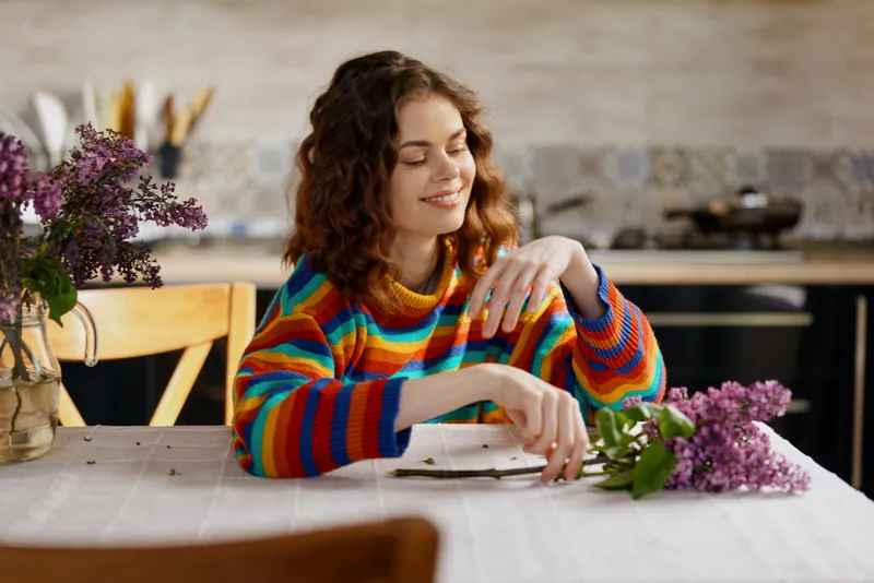 Woman with curly hair in a colorful striped sweater sits at a home kitchen table arranging flowers, smiling softly. Indoor lifestyle scene showcasing creativity, relaxation, and daily beauty.